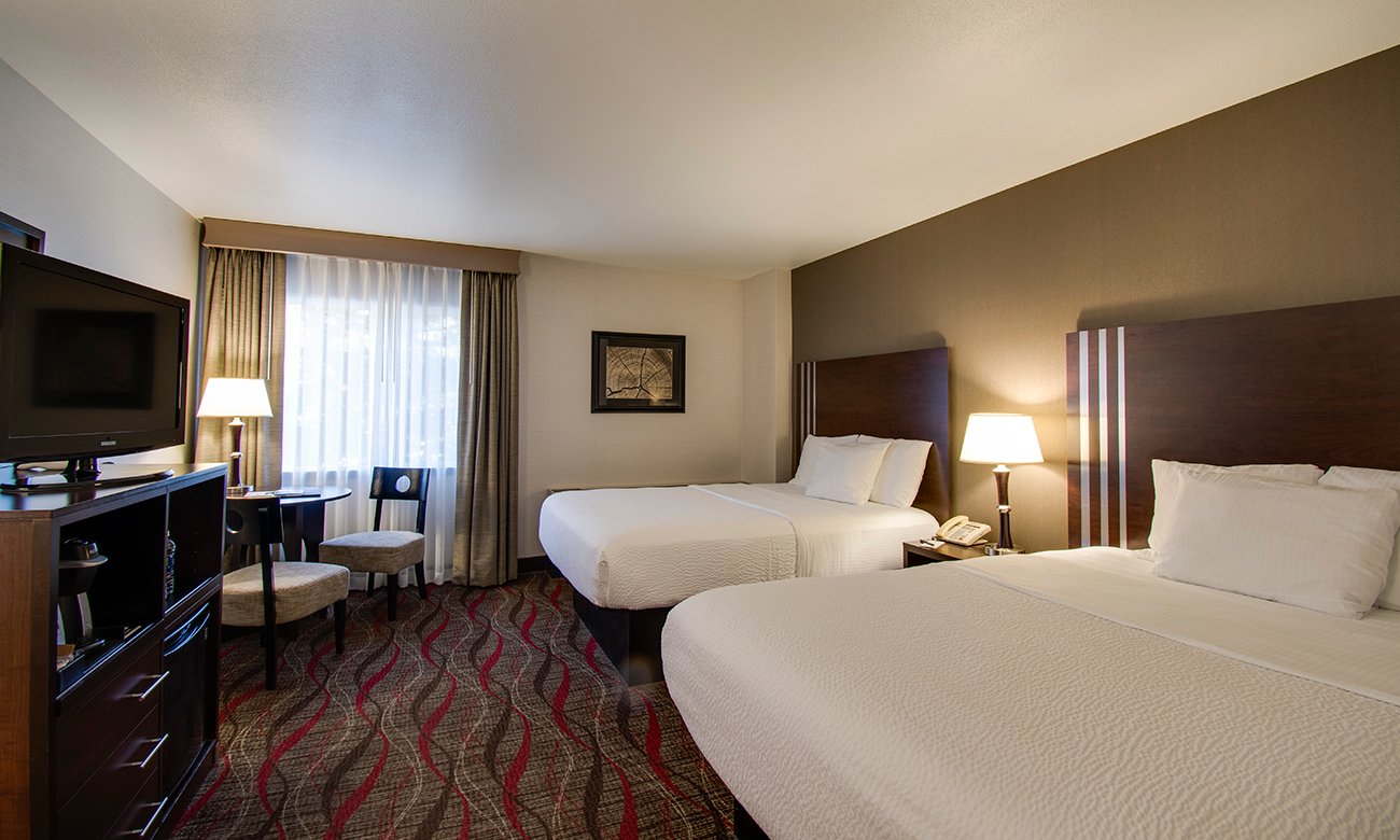 This two bed, west queen hotel room shows the room amenities, including TV, coffee maker, Takelma coffee, seating, a table, and comfortable mattresses.